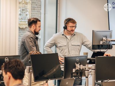 Two colleagues discussing work at a standing desk in a bright, modern office with others working in the background.
