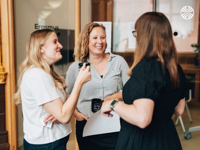 Group of white women in a conversation, smiling, standing in the office hallway.