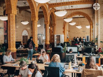 Spacious office with long rows of desks and people working on computers under high, arched ceilings.