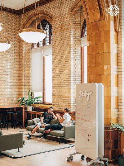 Two white men sitting on a sofa in discussion in an open hallway with arched ceilings.