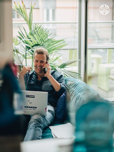 Person relaxing on a sofa taking a phone call, with a Kraken sticker on their laptop and tropical plants nearby.