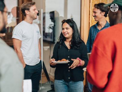 Group of colleagues, including South Asian and white team members, laughing and holding snacks in a casual setting.