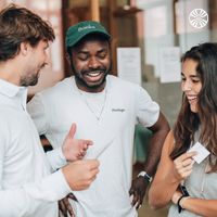 A Black man and a white man talk with a South Asian woman, smiling, near a table in a spacious workspace.