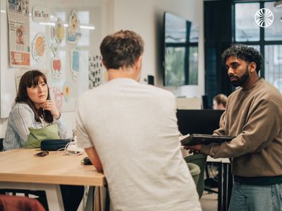Three colleagues having an informal meeting at a table, with colorful artwork posted on the wall behind them.