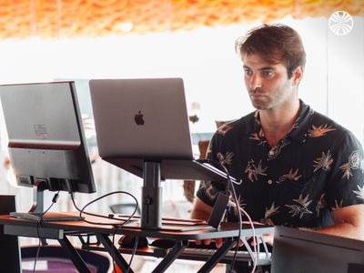 A white man focused on working at a standing desk with a laptop and monitor, in a brightly lit office space with an orange ceiling.