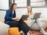 Two white women work together on laptops while seated comfortably on colourful beanbags in an office.