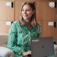 A smiling woman in a green dress sits with a laptop, working in a modern office with wooden lockers in the background.