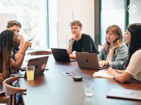 A Black woman leads a discussion at a conference table with three colleagues, two white men and an Asian woman.