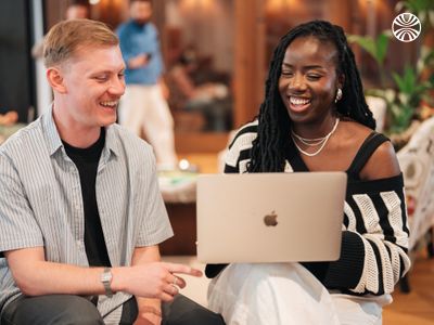 A white man and a Black woman smiling and discussing over a laptop in a vibrant office environment.