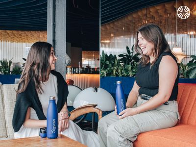 2 white women seated and laughing indoors, each holding a Jigsaw branded bottle.