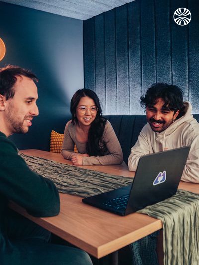 Three team members collaborating over a laptop in a dimly lit restaurant booth, sharing a relaxed work discussion.