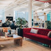 Bright open office featuring a lounge area with red velvet sofa, grey sofa, and games atop a wooden coffee table. Workstations are positioned behind the lounge area.