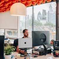 Man wearing glasses working at a desk with multiple monitors, office with cityscape views.