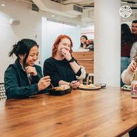 Colleagues sharing lunch and laughter around a wooden table, enjoying takeout meals and beverages in a bright office.