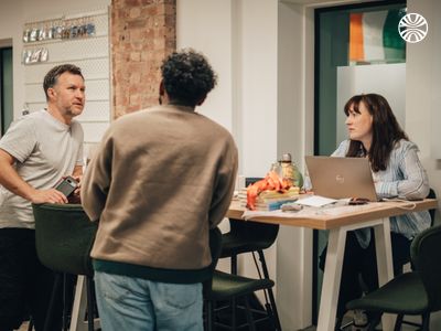 Three colleagues gathered around a laptop during a casual meeting in an industrial-style office space.