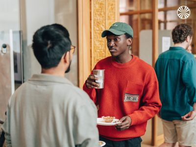 Black man holding a drink and a plate, chatting with a South Asian colleague in a casual office setting.