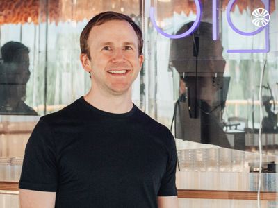 A smiling white man in a black t-shirt standing in front of a glass wall with the partial reflection of a neon sign and office equipment.