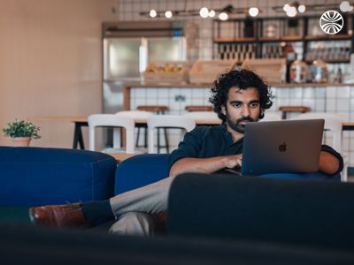 A man with curly hair and beard works on a laptop in a modern office setting with a casual seating area and kitchen in the background.