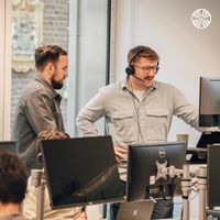 Two colleagues discussing work at a standing desk in a bright, modern office with others working in the background.