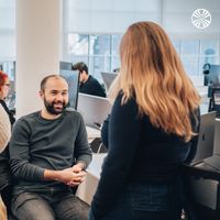 Three team members in conversation. One is seated at their desk facing the other two.