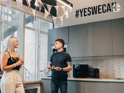 Two colleagues chatting near a "Yes We Can" sign in an office kitchen.