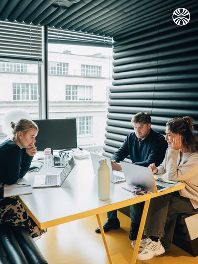 Team meeting with 3 people working on laptops in a modern office space with large windows.