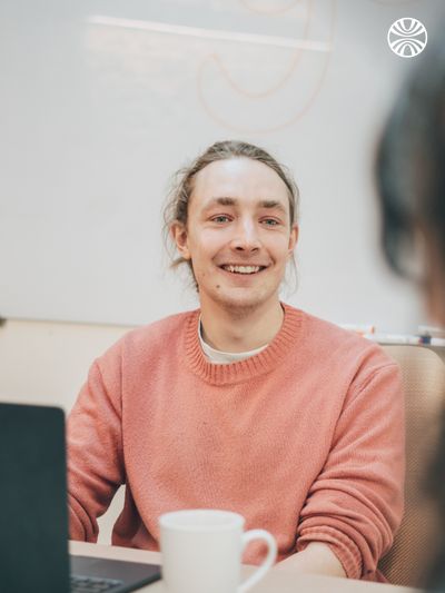 Employee in a coral sweater at their desk with coffee cup, engaged in friendly conversation.