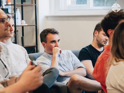 White colleagues seated on a grey couch during a meeting, one person in thoughtful pose with hand near mouth.