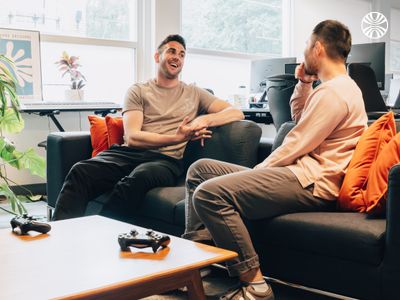 Two white male colleagues laughing and chatting together on a couch in the office.