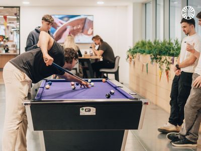 Four colleagues playing pool in a break area, with others working casually in the background.