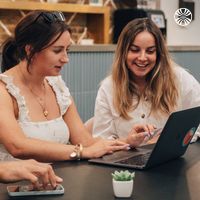 Two women collaborating at a laptop, sitting at a table in a relaxed office environment.