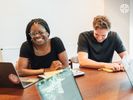 A Black woman laughing with a white male colleague in a meeting around a conference table.