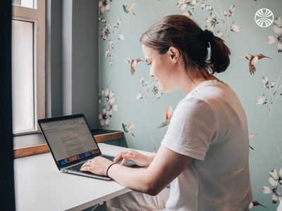 White woman seated, working on a laptop against a floral wallpaper background.