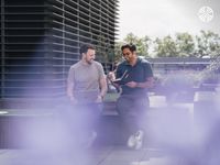 Two men sit outside with laptops, discussing notes from a notebook, against a building facade.