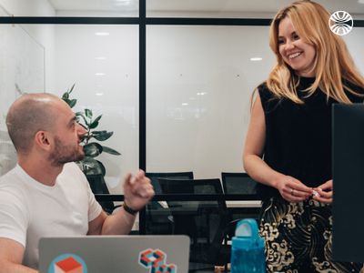 White man speaking to a white woman standing, both smiling in a casual office conversation.