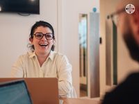 A woman in glasses and a white shirt smiles while talking to a colleague in an office setting.