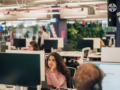 Open office space with multiple workstations and indoor plants, showing team members focused on their work.