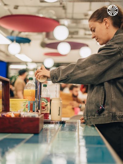 Person refilling drink from the office kitchen counter with colorful food options displayed.