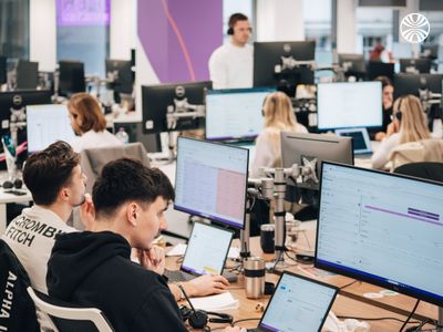 A large group working at individual computer workstations in an open-plan office with purple accent walls.