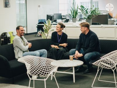 3 people having an animated discussion while seated on a sofa around a low table, with office plants in the background.