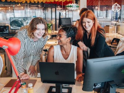 2 white women and one Black woman collaborating at a desk with a laptop in an office.