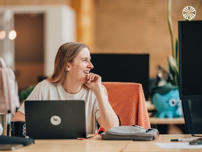 White woman smiling and looking to her side while working at a laptop in a bright office.