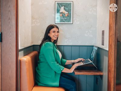 A multiracial woman working on a laptop in an office phone booth smiles at the camera.