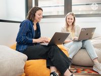 Two white women work together on laptops while seated comfortably on colourful beanbags in an office.