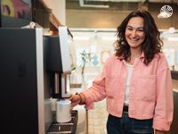 A smiling white woman using a coffee machine in an office kitchen.