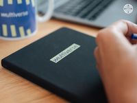 Close-up of a multiverse-branded black notebook and a blurred laptop in the background.