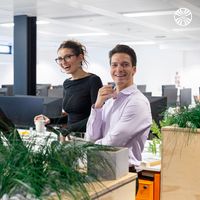 Two team members standing and sharing a cheerful moment at their desk, with office plants in the foreground.