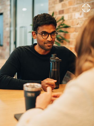 Person wearing glasses and a black sweater engaged in conversation over coffee, with a reusable water bottle on a wooden table.