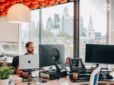 Man wearing glasses working at a desk with multiple monitors, office with cityscape views.