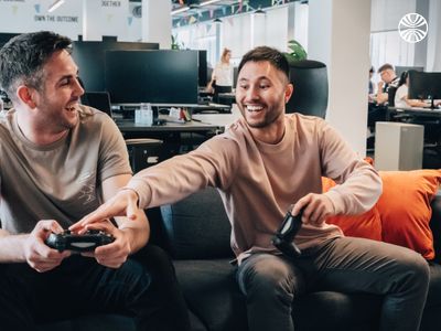 Two white male colleagues laughing while gaming on an orange-cushioned couch.